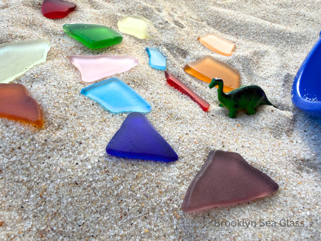 Bright tumbled glass in rainbow colors with a small dinosaur toy in sand.